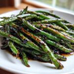 Close-up of bright green beans coated in a dark, glossy chinese green beans with garlic sauce on a white plate.