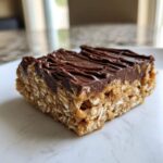 A single, square homemade protein bars slice with an oat base and thick chocolate topping drizzled with more chocolate.