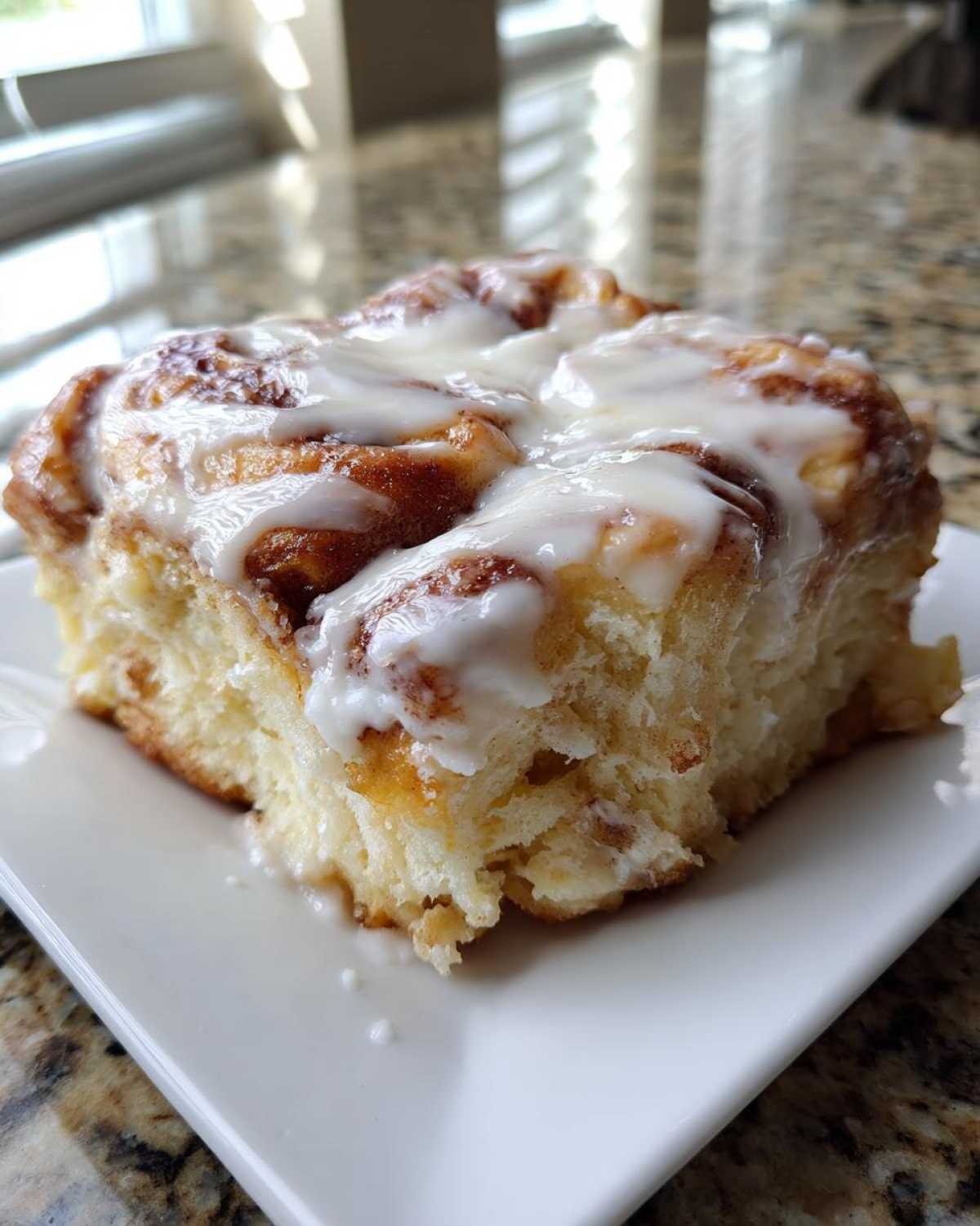 A close-up of a fluffy, square slice of cinnamon roll casserole topped with thick white icing.