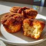 Three cinnamon sugar coated french toast muffins on a white plate, one is broken open showing the soft interior.