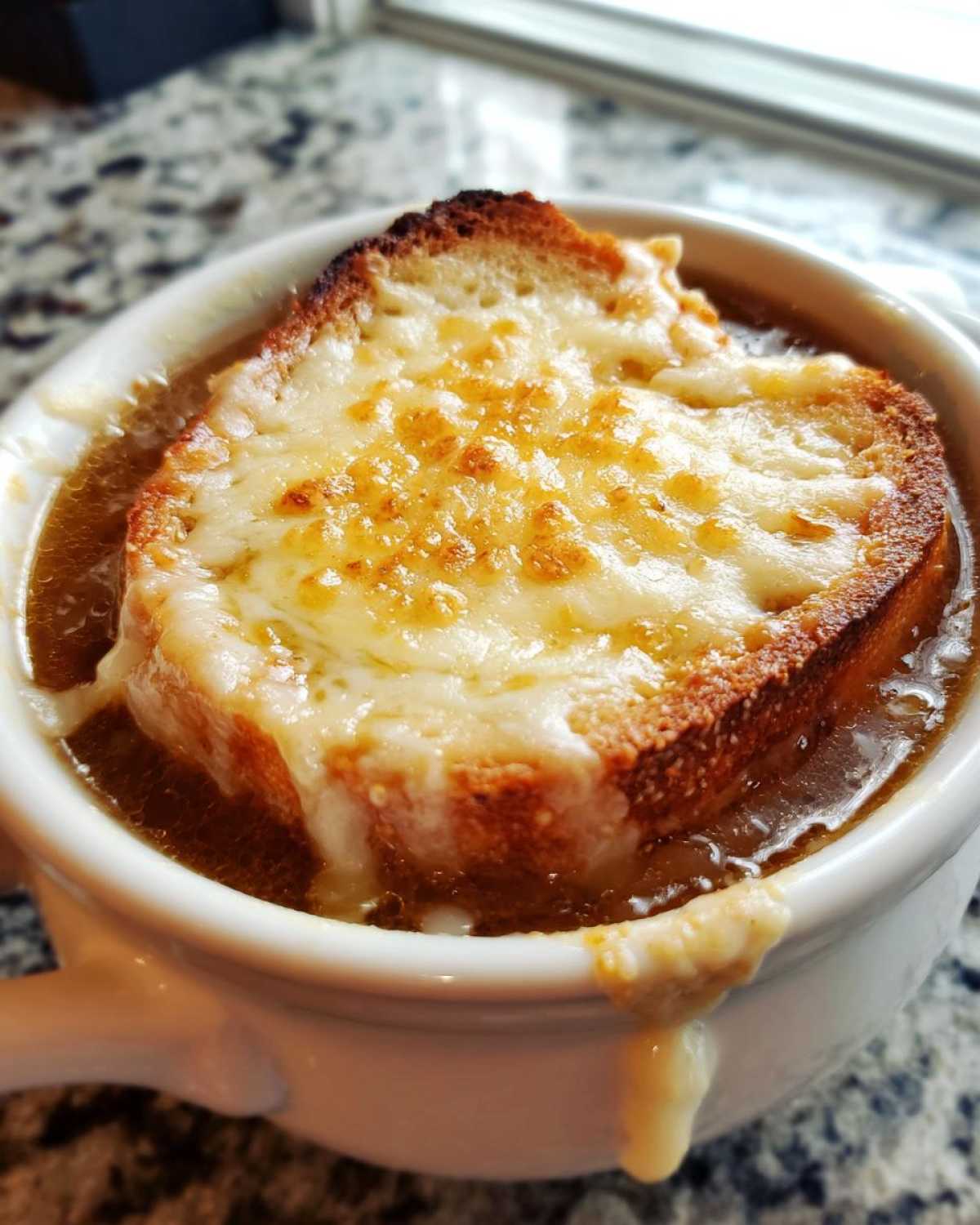 Close-up of a white crock of rich french onion soup topped with a toasted crouton and bubbly, melted Gruyère cheese.