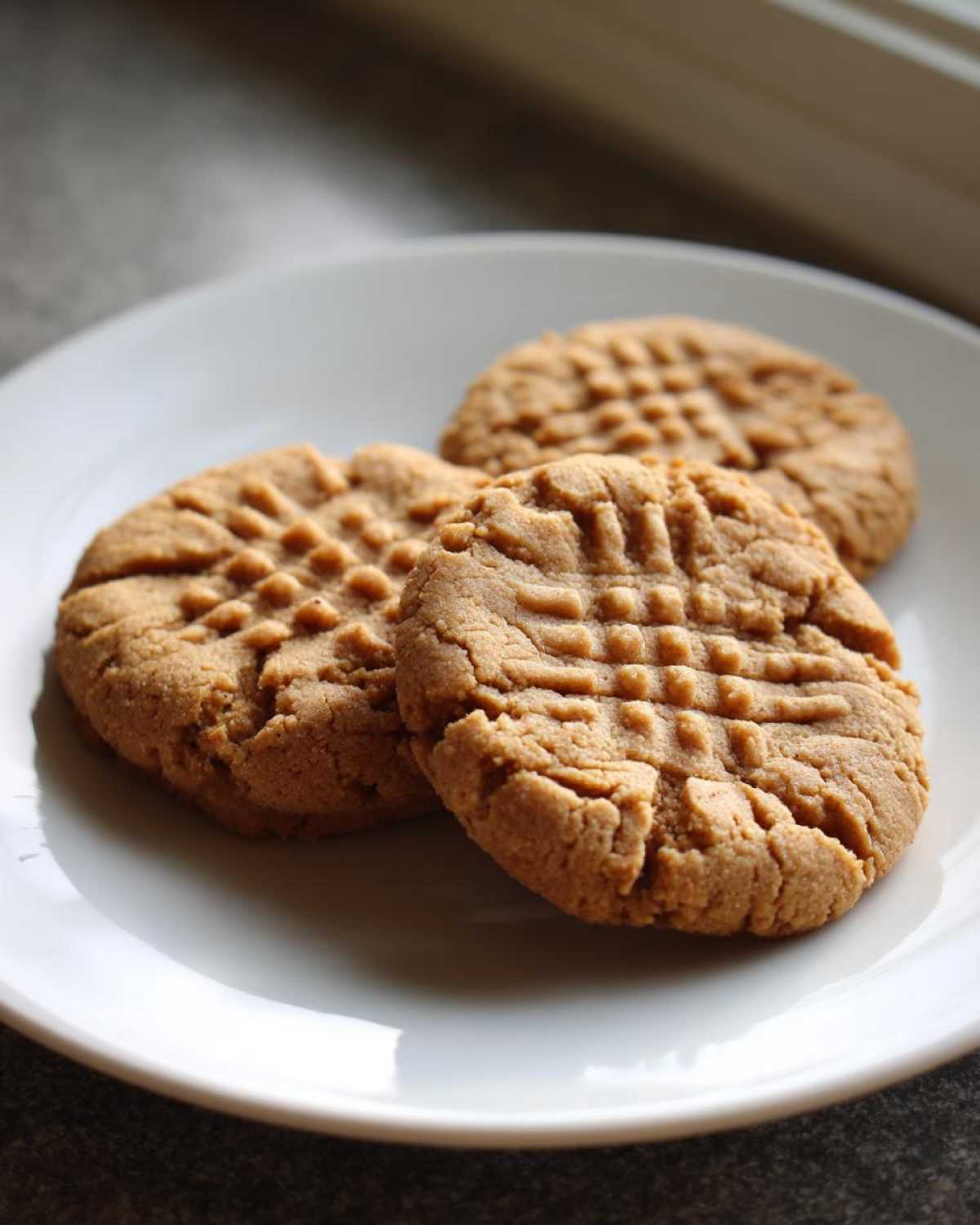 Three freshly baked peanut butter cookies with classic fork crosshatch marks served on a white plate.
