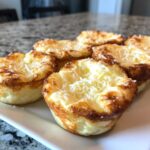 Close-up of six golden brown cottage cheese egg cups starbucks hack, topped with shredded cheese, on a white plate.