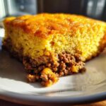 A close-up of a slice of cowboy cornbread casserole showing the thick, golden cornbread top layer over a savory ground meat filling.