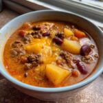 A close-up of a bowl of rich, orange cowboy soup topped with melted cheddar cheese, ground beef, potatoes, and kidney beans.