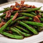 A close-up of glossy, bright green beans coated in a dark sauce and topped with crispy bacon pieces.
