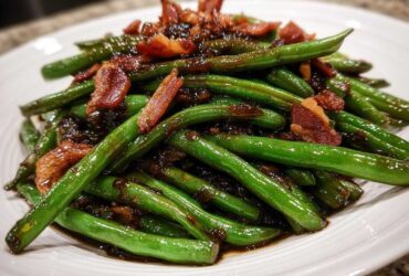 A close-up of glossy, bright green beans coated in a dark sauce and topped with crispy bacon pieces.