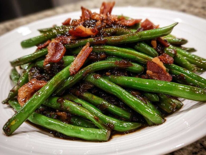 A close-up of glossy, bright green beans coated in a dark sauce and topped with crispy bacon pieces.
