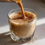 Close-up of creamy coquito being poured into a glass, topped with cinnamon.