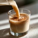 Close-up of creamy coquito being poured into a glass already dusted with cinnamon.
