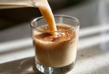 Close-up of creamy coquito being poured into a glass already dusted with cinnamon.