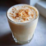 Close-up of a glass filled with creamy coquito recipe, topped with frothy foam and sprinkled cinnamon.