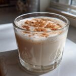 A glass filled with creamy, frothy coquito recipe, dusted generously with ground cinnamon.