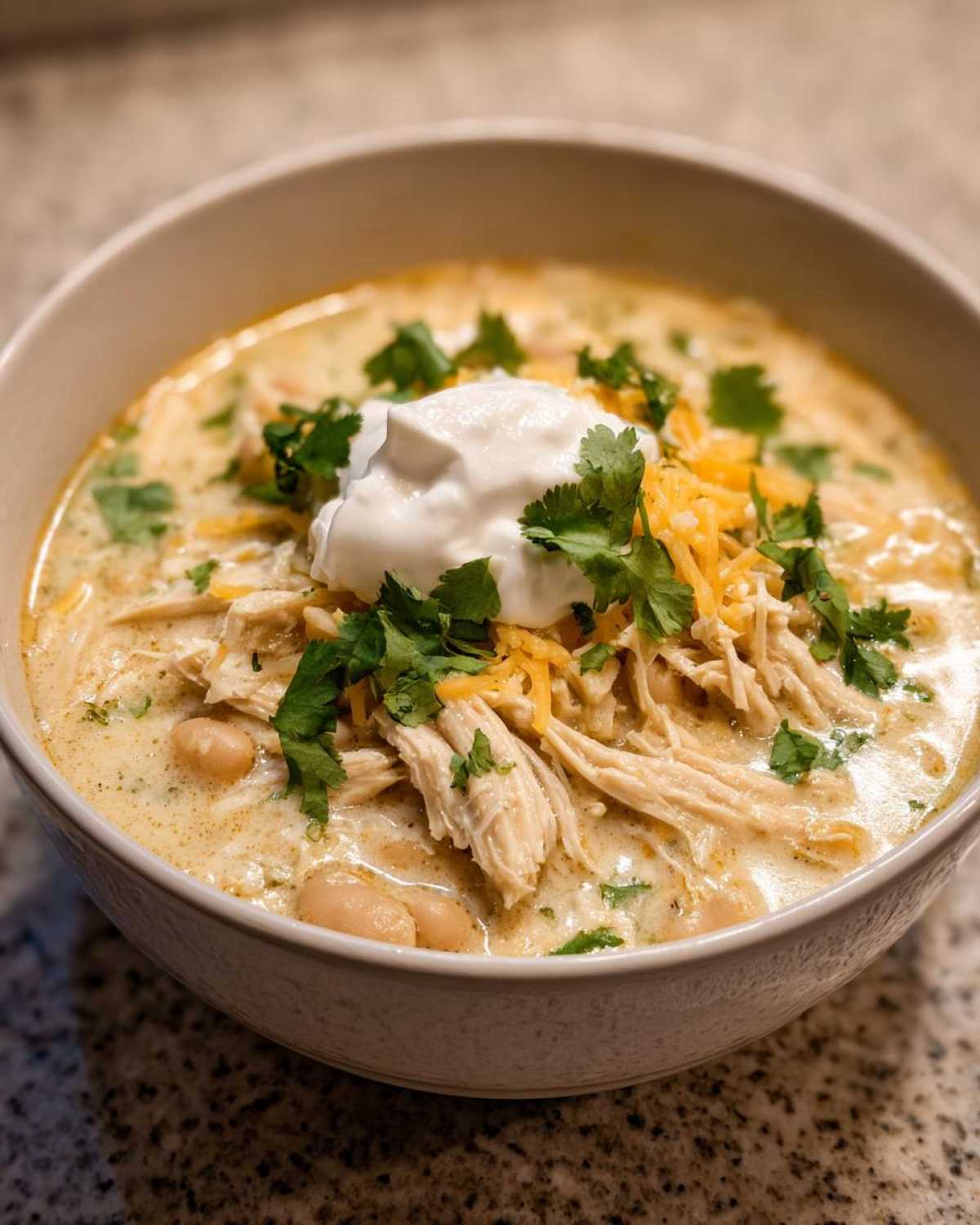 A close-up of a bowl of creamy crockpot white chicken chili topped with shredded chicken, sour cream, cheese, and cilantro.