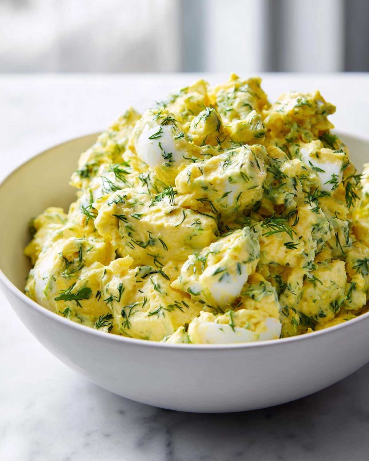 A close-up of a creamy, bright yellow egg salad recipe mixed with fresh dill, served in a white bowl.