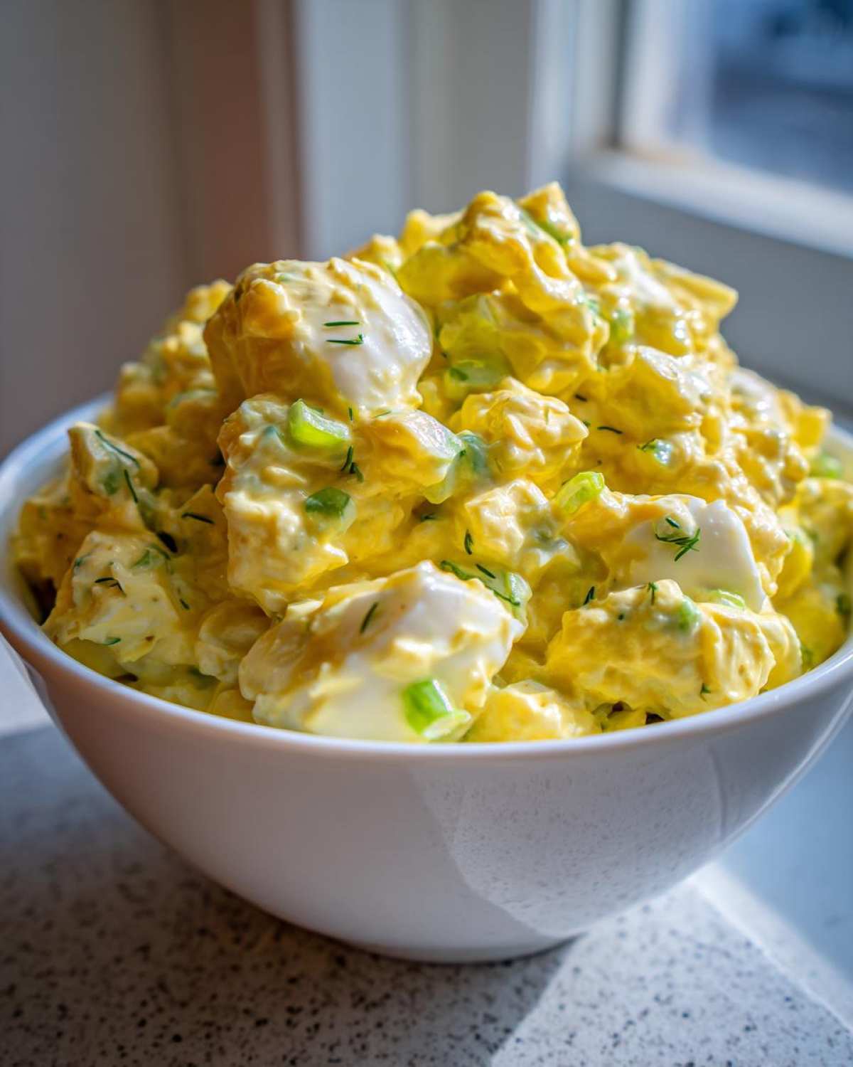 Close-up of a bowl filled with creamy, yellow egg salad recipe, featuring chunks of egg white and green herbs.