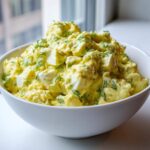 A large white bowl filled with creamy, yellow egg salad recipe, garnished with fresh dill.