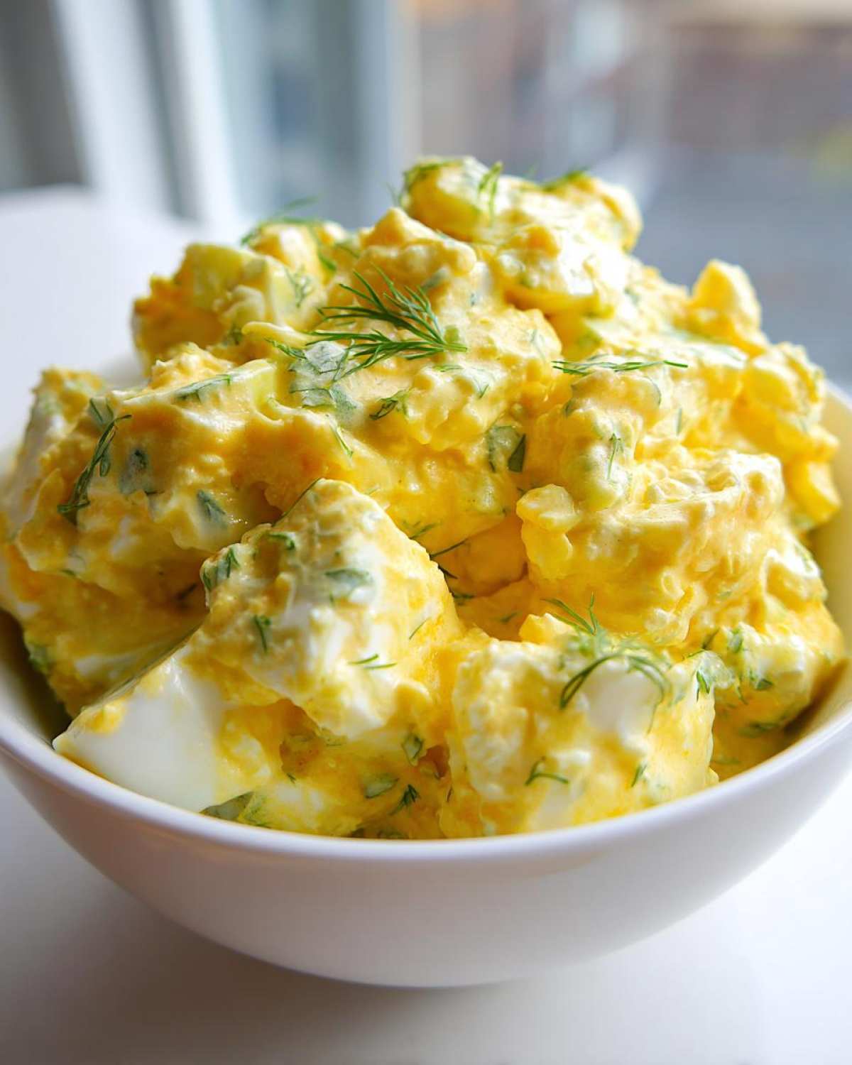 A close-up of creamy, chunky egg salad recipe garnished with fresh dill in a white bowl.