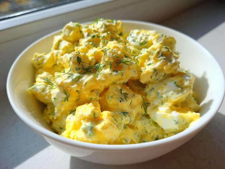 Close-up of a bright yellow, creamy egg salad recipe garnished with fresh dill in a white bowl.