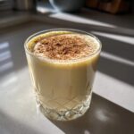 A glass of creamy, pale yellow eggnog topped generously with ground cinnamon, sitting on a bright countertop.