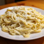 A plate of fettuccine pasta generously coated in creamy homemade alfredo sauce and topped with grated Parmesan cheese and black pepper.