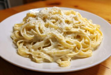 A plate of fettuccine pasta generously coated in creamy homemade alfredo sauce and topped with grated Parmesan cheese and black pepper.
