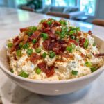 A white bowl filled with creamy Million Dollar Dip, generously topped with crumbled bacon and fresh green onions.