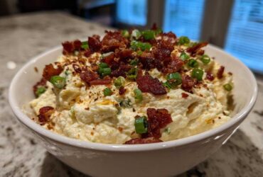 A close-up of creamy Million Dollar Dip served in a white bowl, generously topped with crumbled bacon and sliced green onions.