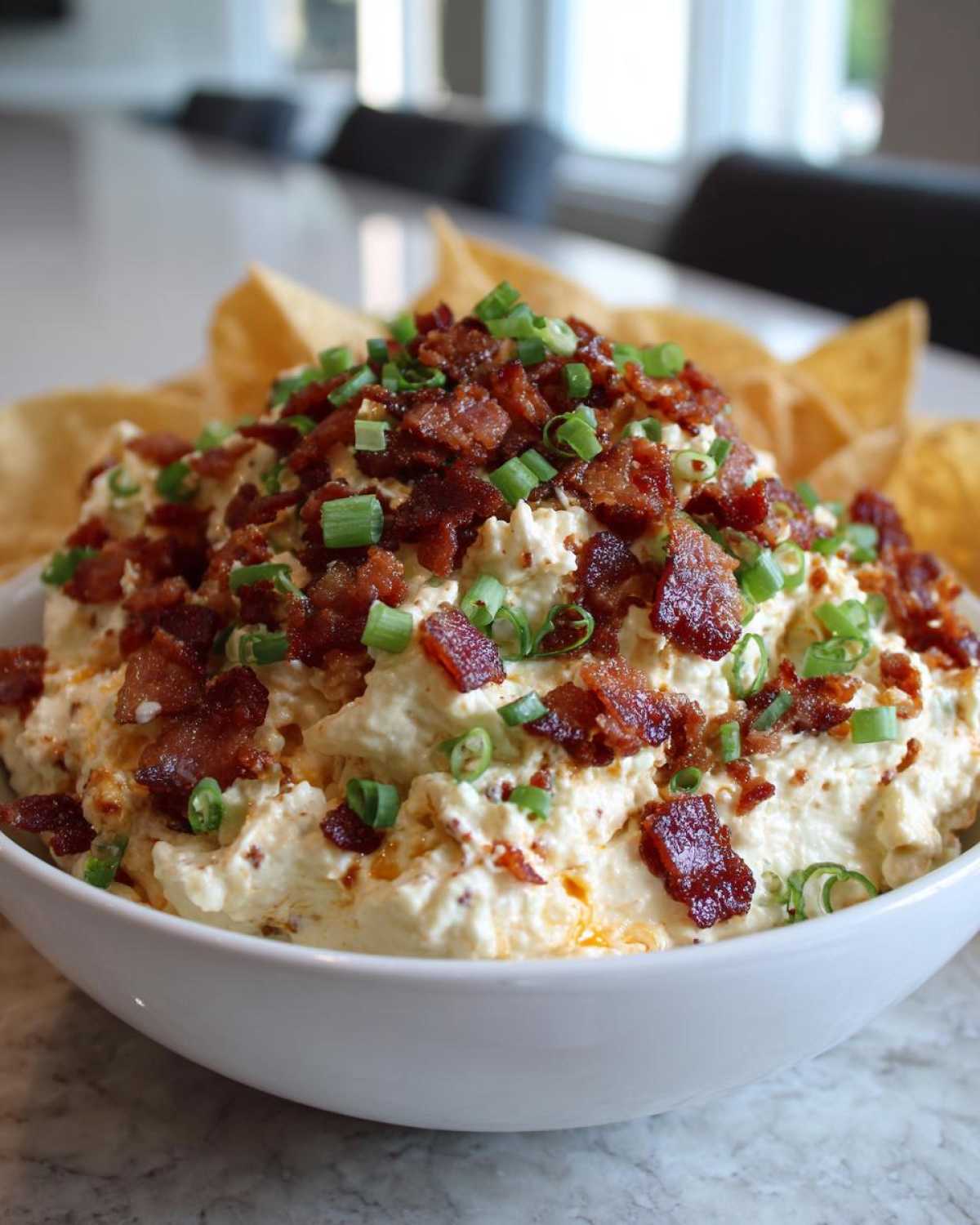 A creamy bowl of million dollar dip generously topped with crispy bacon bits and sliced green onions, served with tortilla chips.