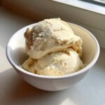 Two scoops of creamy, pale yellow protein ice cream served in a small white bowl near a window.