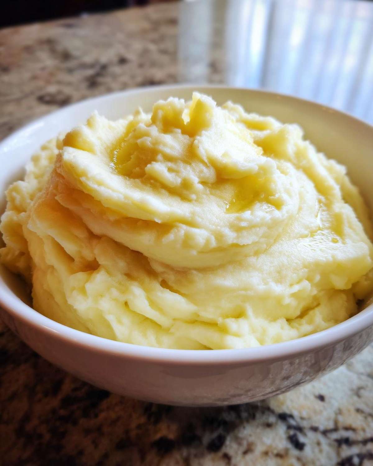 A close-up of fluffy roasted garlic mashed potatoes, swirled high in a white bowl with melted butter pooling on top.