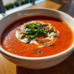 A close-up of a bowl of rich, creamy tomato bisque, swirled with cream and topped with fresh basil.