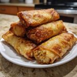 A stack of golden brown, crispy fried egg roll appetizers piled on a white plate, one showing the filling.