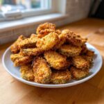 A large pile of golden brown, crispy Fried Pickles stacked high on a white plate on a wooden surface.