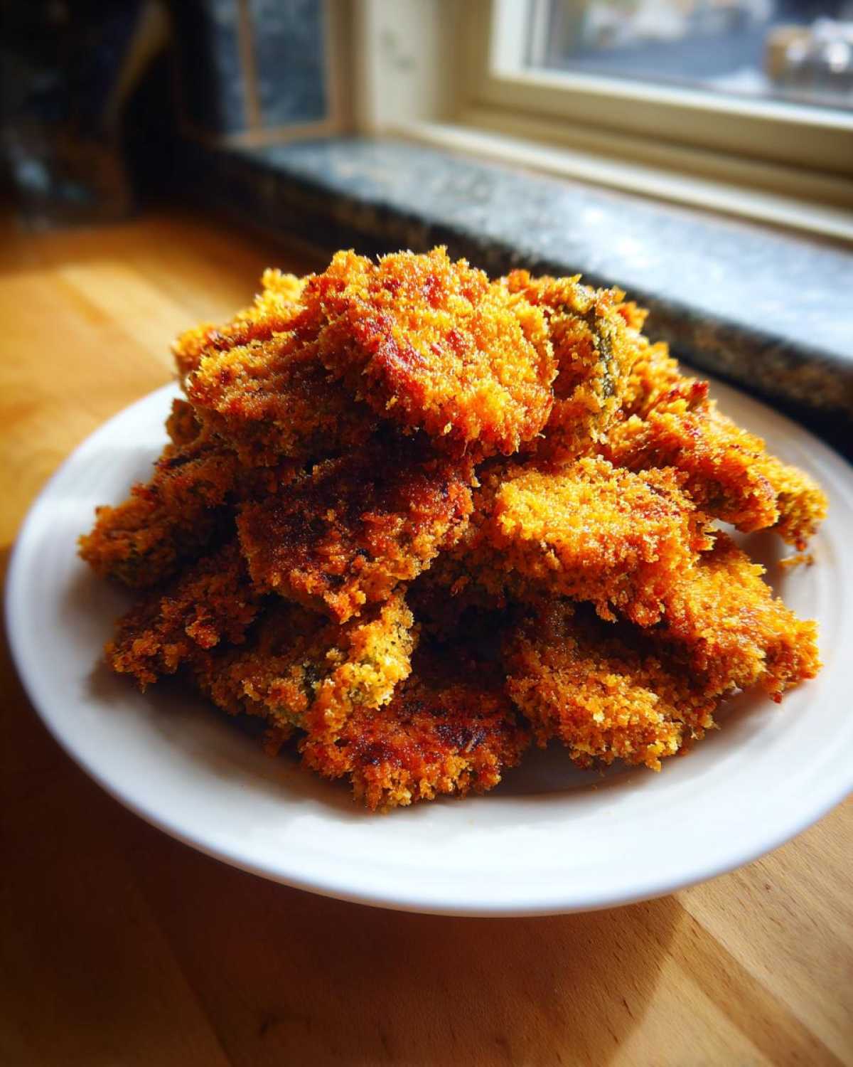 A tall pile of golden brown, crunchy Fried Pickles stacked high on a white plate indoors.