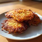 Four perfectly crispy, golden-brown potato latkes stacked on a light blue plate, illuminated by sunlight.