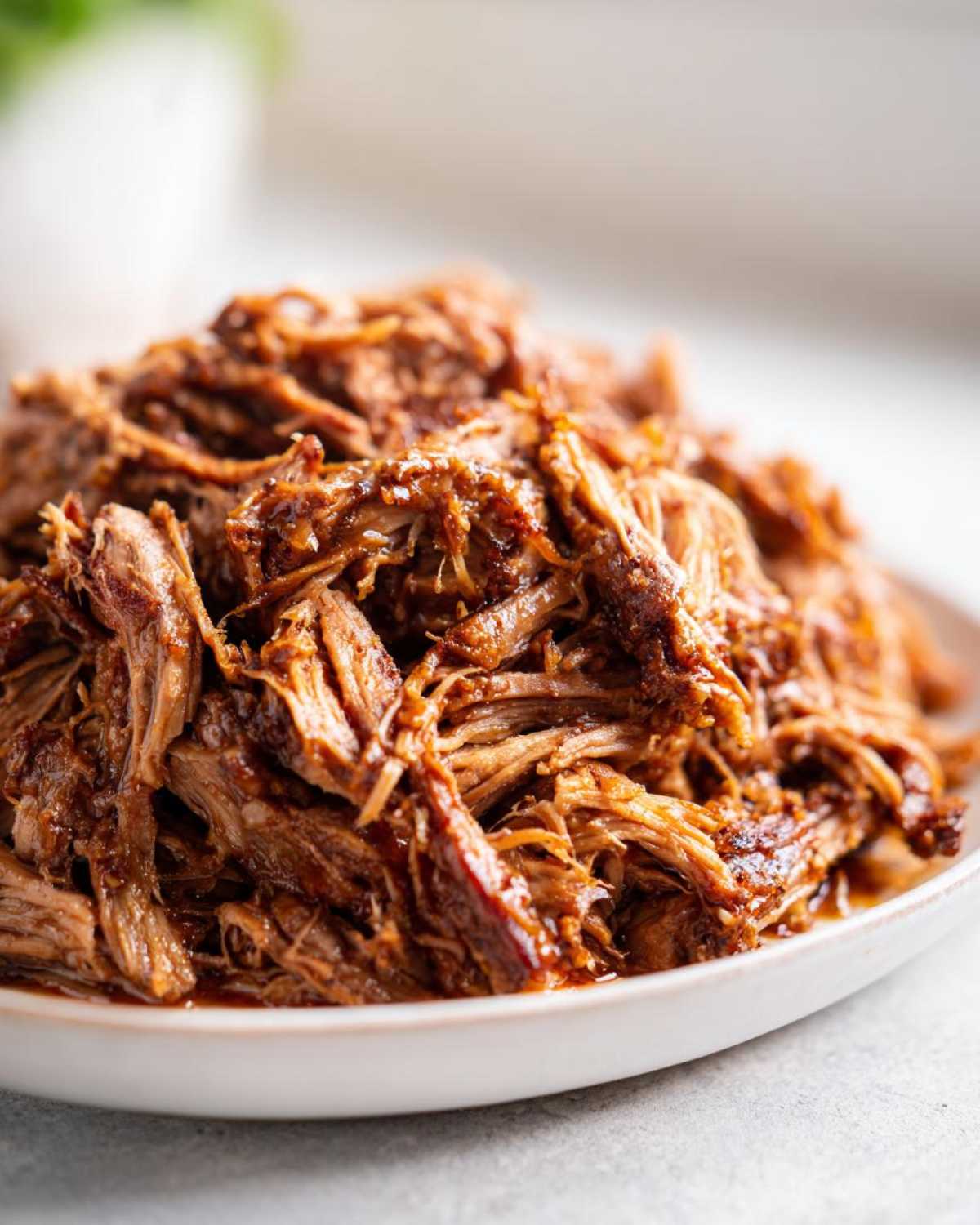 Close-up of tender, shredded crock pot pulled pork coated in a rich, dark barbecue sauce on a white plate.