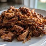 Close-up of tender, saucy crock pot pulled pork shredded and piled high on a white plate.