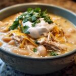 A close-up of a bowl of creamy crockpot white chicken chili topped with sour cream, shredded cheese, and fresh cilantro.