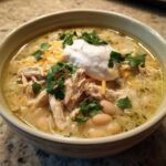 A close-up of a bowl of creamy crockpot white chicken chili topped with shredded chicken, white beans, sour cream, cheese, and cilantro.