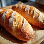 Two golden-brown, crusty loaves of homemade french bread resting on parchment paper, showing beautiful scoring.