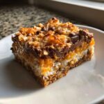 A single, rich square of magic cookie bars showing layers of graham cracker crust, coconut, caramel, and chocolate chips.