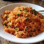 A close-up shot of fluffy, reddish-orange spanish rice mixed with bright green peas and diced carrots in a white bowl.