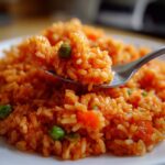 A spoonful of bright orange spanish rice mixed with green peas being lifted from a white plate.
