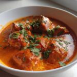 Close-up of rich, orange-red butter chicken pieces garnished with fresh cilantro in a white bowl.