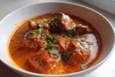 Close-up of rich, orange-red butter chicken pieces garnished with fresh cilantro in a white bowl.