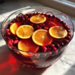 Close-up of a vibrant thanksgiving punch garnished with floating orange slices and fresh cranberries in a clear glass bowl.