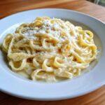 A close-up of fettuccine pasta coated in rich homemade alfredo sauce, topped with grated Parmesan and black pepper.