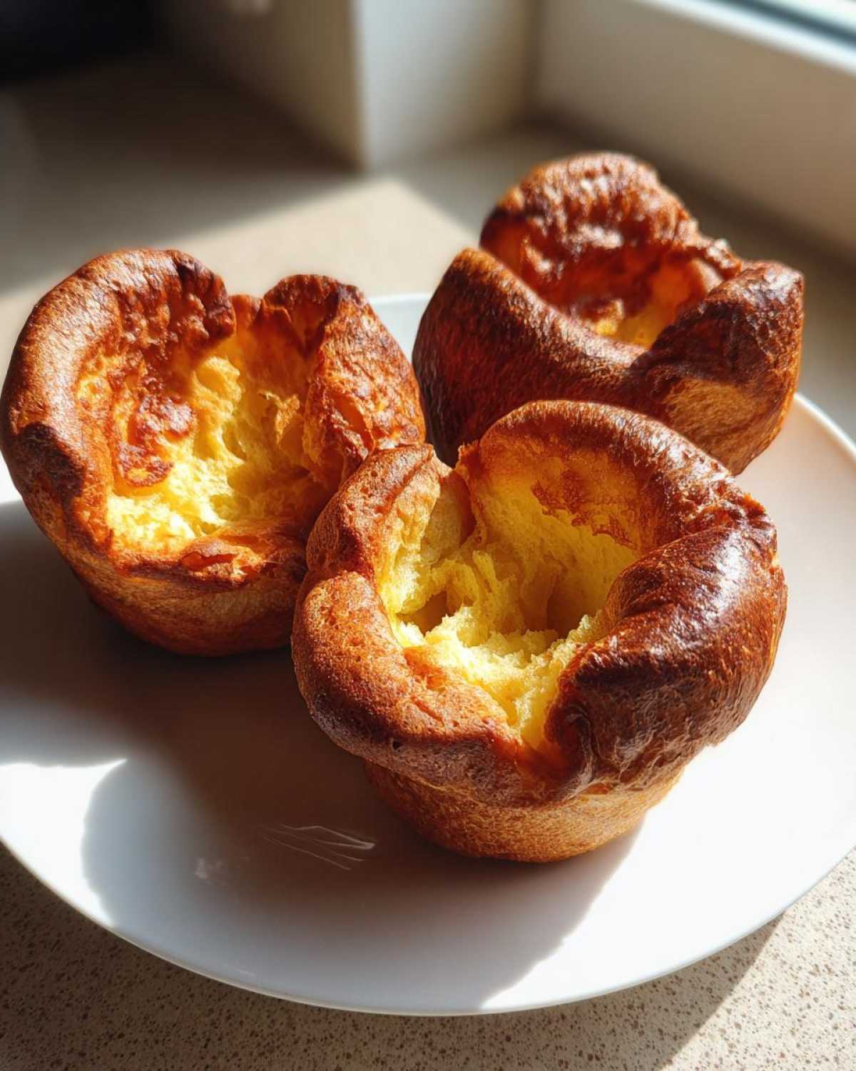Three golden brown, perfectly risen yorkshire pudding cups sitting on a white plate, catching bright sunlight.