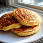 A stack of three golden brown, fluffy almond flour pancakes on a white plate near a window.